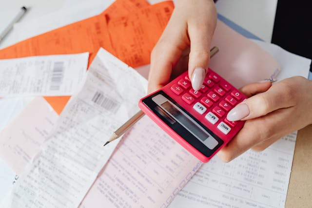 Person holding a calculator and pencil abouve a table full of receipts